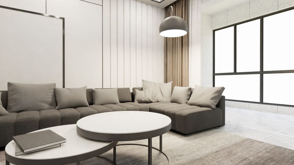 A modern living room with a large, L-shaped grey fabric sofa topped with multiple matching cushions, positioned against a white paneled wall. In front of the sofa, there is a round white coffee table