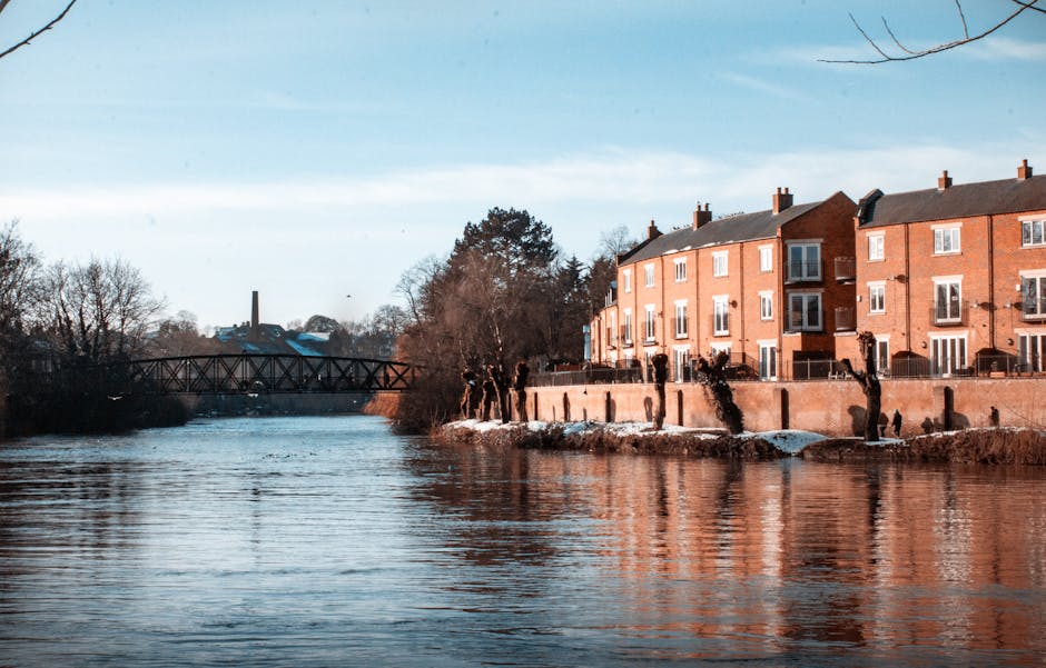A riverside residential area with red brick apartment buildings lining the bank of a calm river. The buildings feature large windows and balconies, with some having fencing or railings. A stone or con