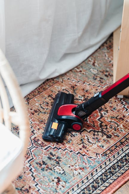 A handheld vacuum cleaner with a red and black design resting on a patterned Oriental-style carpet in a residential room. The carpet features intricate geometric and floral motifs in warm tones of red, orange, and beige. The setting includes a light-colored curtain draped behind the carpet and part of a wooden furniture piece visible nearby. The area appears clean, with no visible dust or debris, indicating a recent surface cleaning or deep cleaning process carried out by Carpet Cleaning SW15. The lighting is natural, highlighting the texture of the carpet and the surface cleanliness, emphasizing professional domestic cleaning and carpet sanitisation.